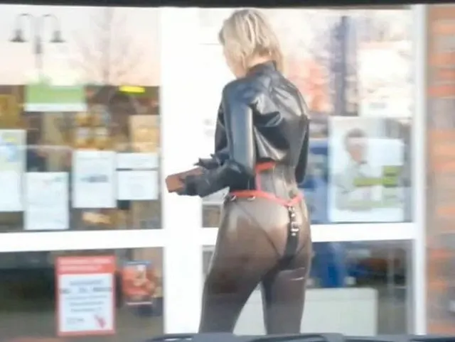 Stark durchbohrte Latexpuppe im transparenten Catsuit Shopping Piercing in der Öffentlichkeit