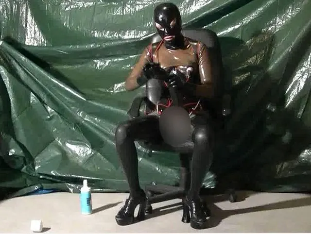 Schweres durchbohrtes Latex-Mädchen in transparenten Catsuit-Nippellöchern, Korsett, Maske masturbiert riesigen Dildo P1